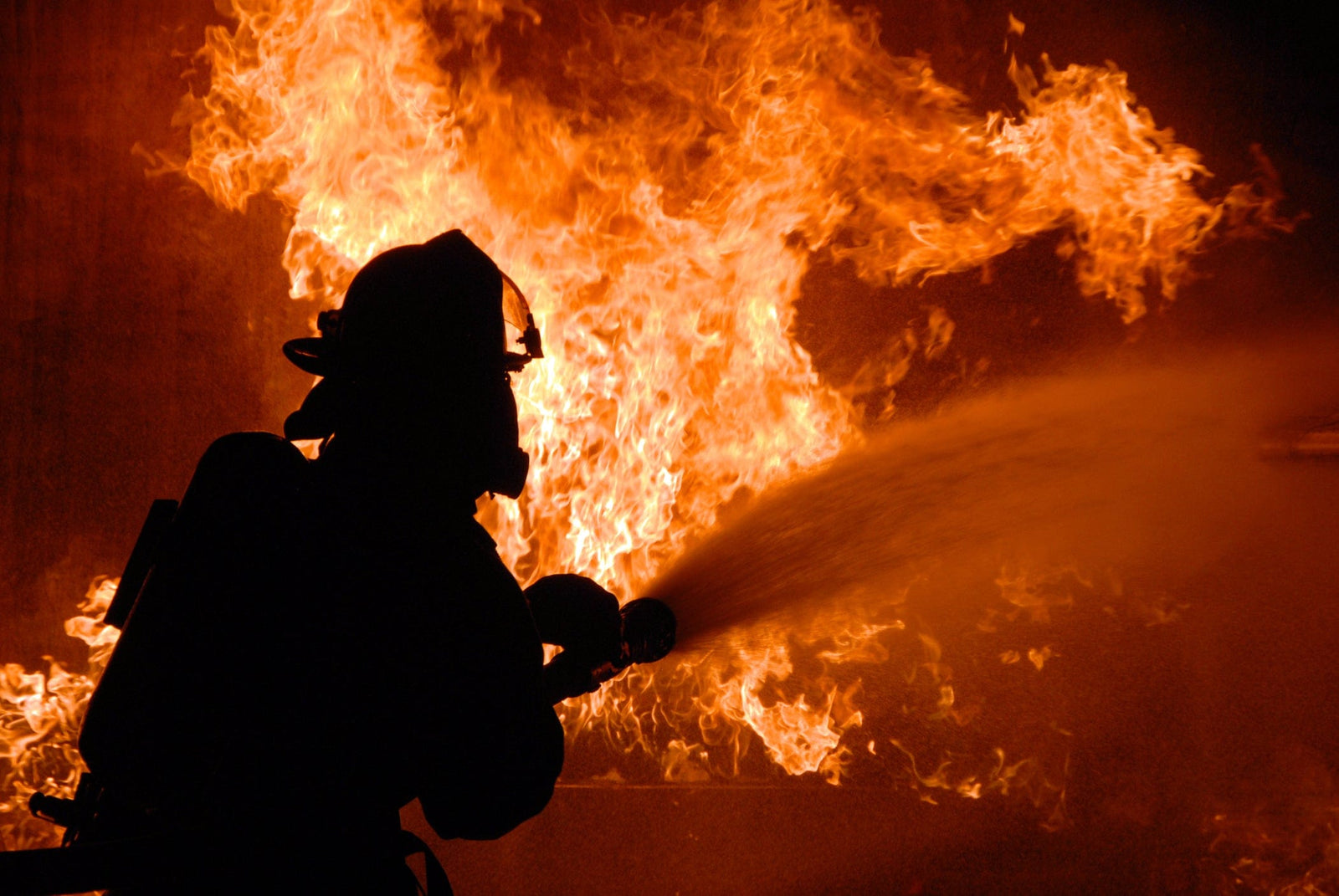 Silhouette of a firefighter spraying water on a large structure fire, surrounded by intense flames and heavy heat during active fire suppression operations.
